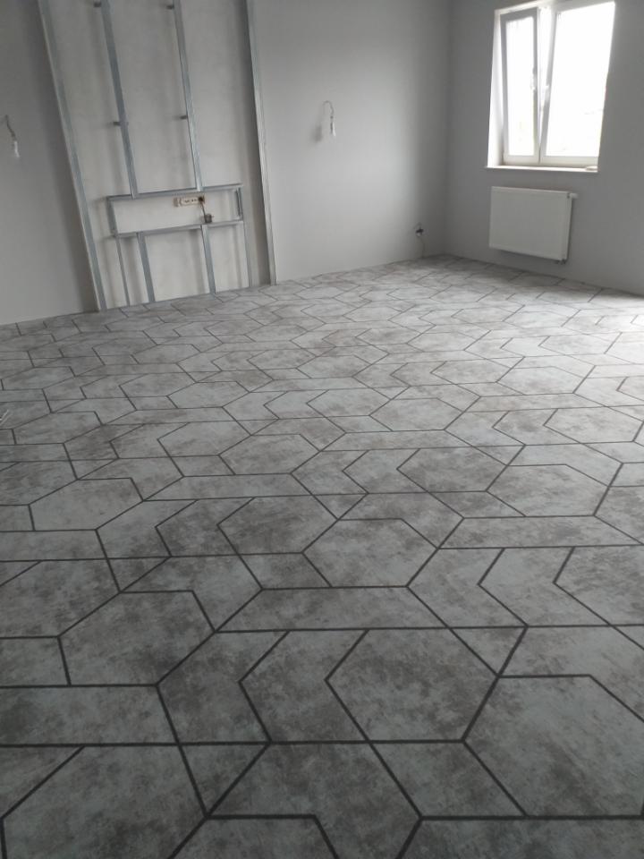 Ukończona podłoga w pokoju z szarymi, heksagonalnymi płytkami imitującymi beton, widoczne przygotowane profile ścienne i grzejnik pod oknem.