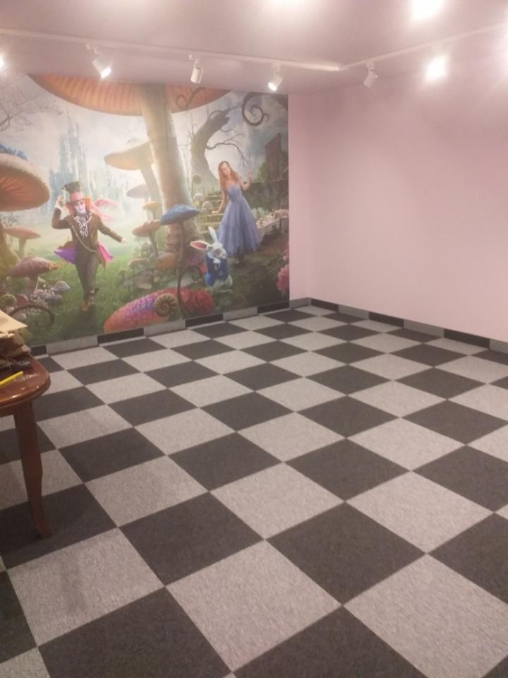 Szachownica z szarej i czarnej wykładziny w pokoju z różowymi ścianami i muralem z Alicji w Krainie Czarów.