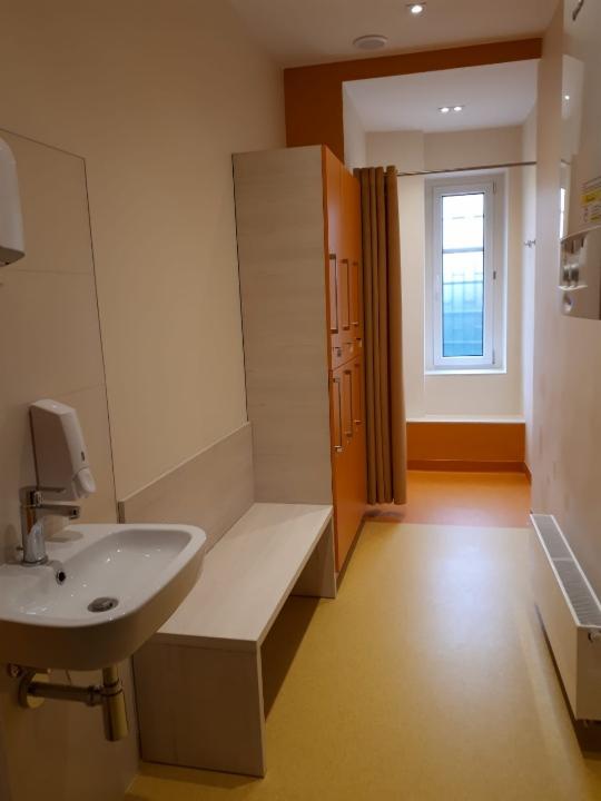 Wnętrze szatni z żółtą podłogą, umywalką, ławką i szafkami, widok na okno z zasłoną.