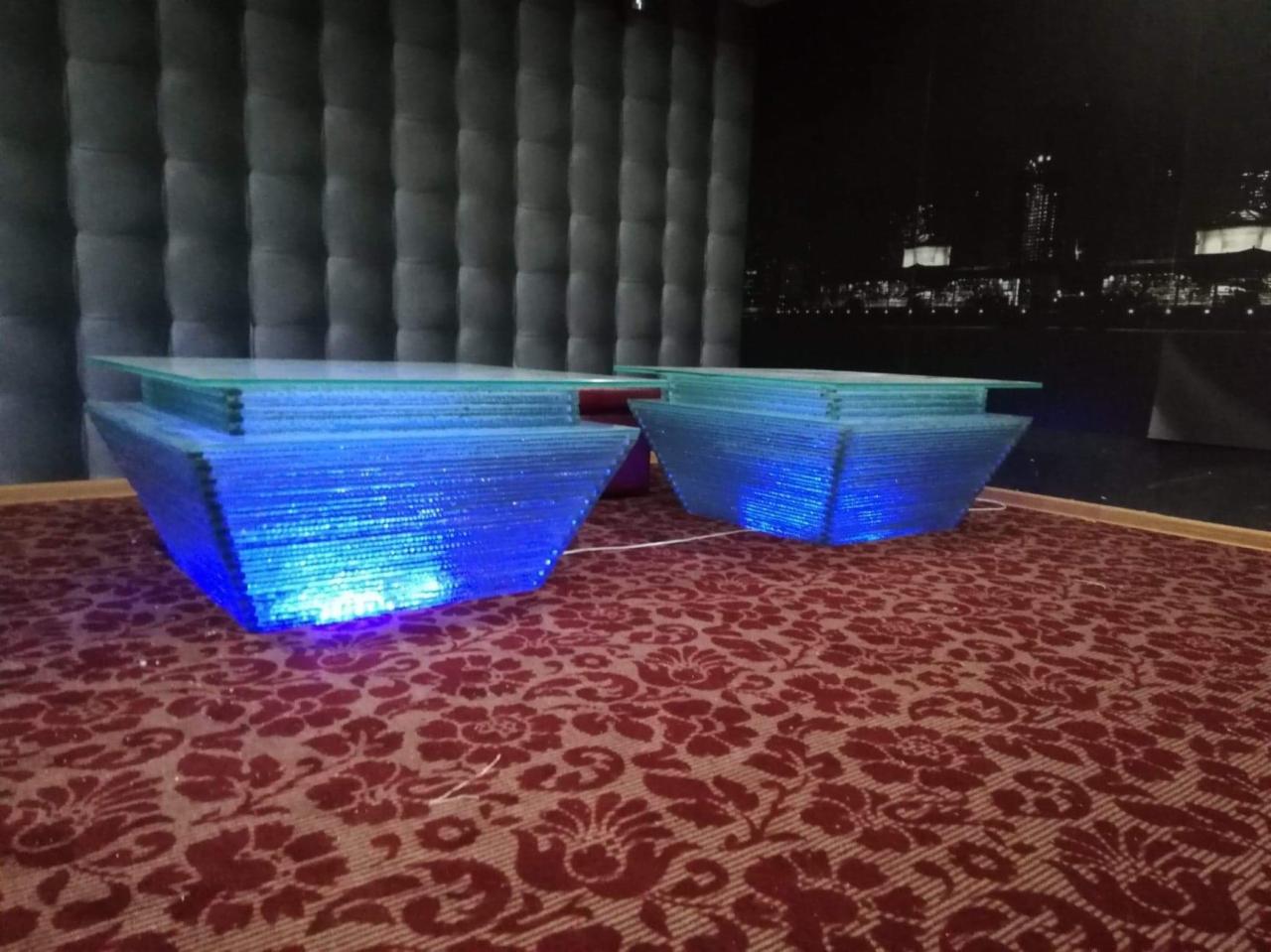 Dwa podświetlane niebieskim światłem stoliki kawowe z przezroczystymi blatami, ustawione na wzorzystej czerwonej wykładzinie, na tle tapicerowanej ściany i panoramy miasta w tle.