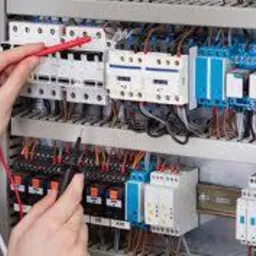 Dłonie elektryka w trakcie podłączania przewodów w rozdzielnicy elektrycznej. Widoczne bezpieczniki, styczniki i inne elementy automatyki. Czerwony przewód w ręce elektryka.