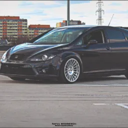 Czarny samochód typu hatchback na betonowym parkingu z budynkami mieszkalnymi w tle, prezentujący zmodyfikowane zawieszenie i felgi.