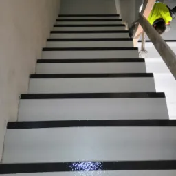 Schody w Krakowie z szarą, świeżo nałożoną powłoką z czarnymi antypoślizgowymi paskami, widoczne stopy pracownika w dolnej części kadru i fragment rusztowania w tle.