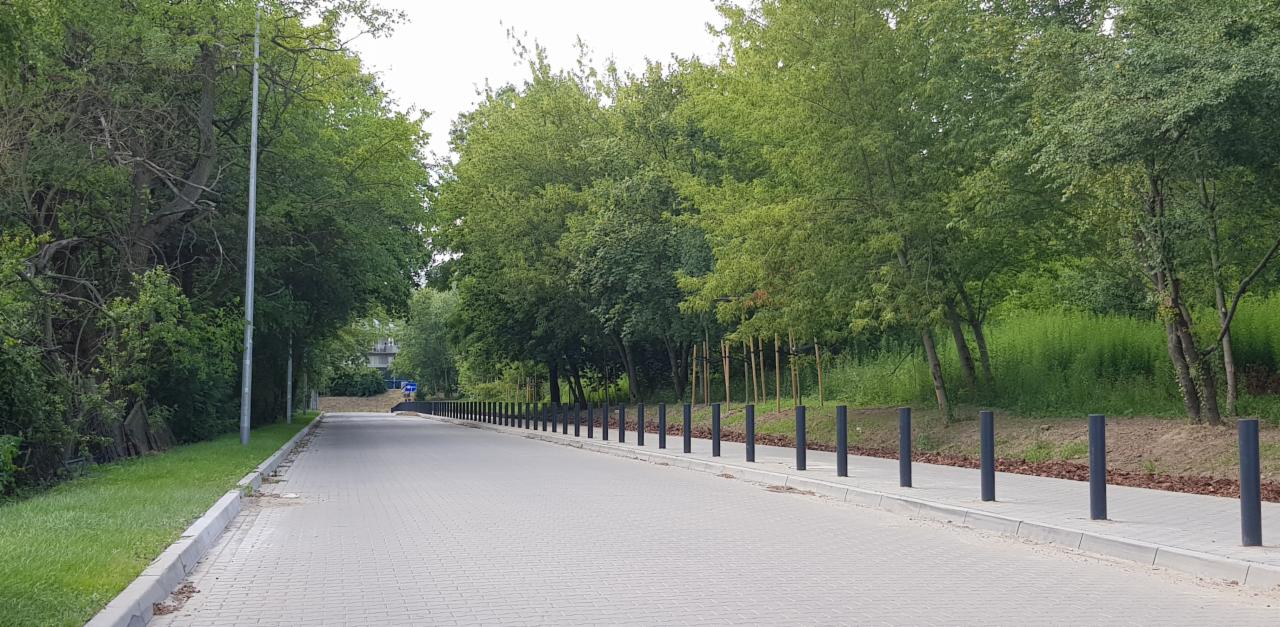 Nowa droga osiedlowa z kostki brukowej, obustronnie zabezpieczona słupkami, otoczona zielenią.