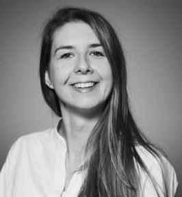 Portret uśmiechniętej kobiety z długimi włosami w czarno-białej tonacji, ubrana w jasną koszulę. Neutralne tło, naturalny wyraz twarzy, delikatne zmarszczki wokół oczu.