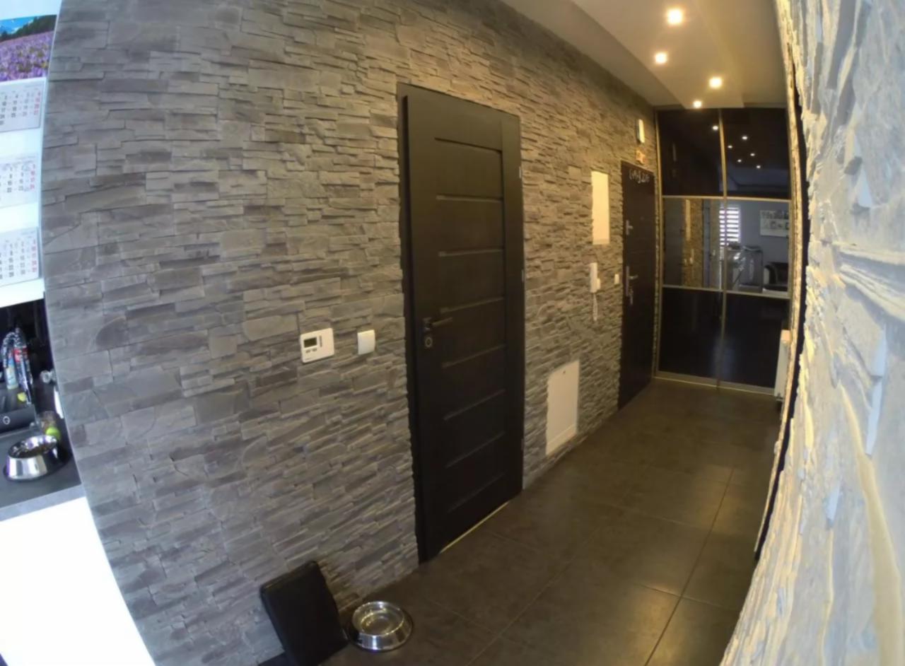 Korytarz wykończony szarą okładziną imitującą kamień, z ciemnymi drzwiami i brązowymi płytkami podłogowymi, widoczna miska dla zwierząt.