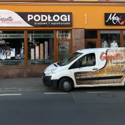 Biała furgonetka z reklamą firmy Geppetto oferującej podłogi drewniane, zaparkowana przed budynkiem z szyldem 'Podłogi krajowe i egzotyczne' oraz wypożyczalnią cykliniarek.