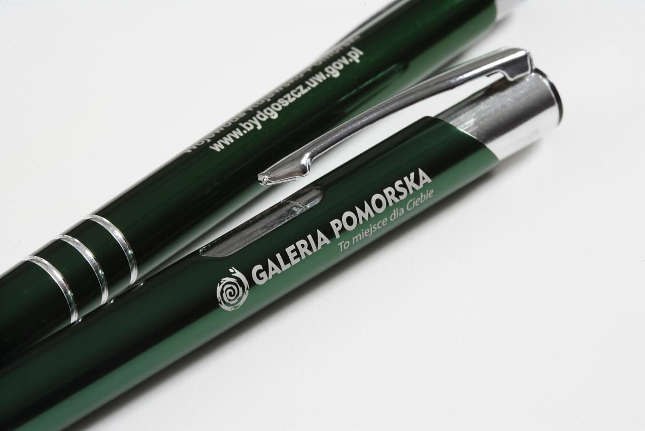Dwa zielone długopisy z nadrukowanym logo 'Galeria Pomorska' i adresem strony internetowej, leżące na białym tle.