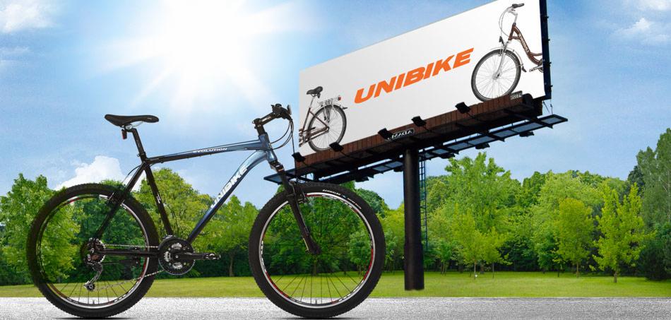 Rower górski marki Unibike na tle billboardu reklamującego rowery tej samej marki, ustawiony na zielonym trawniku z drzewami w tle.