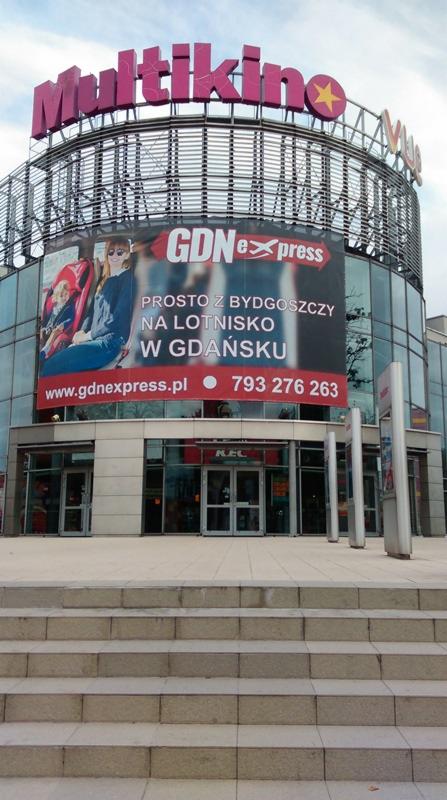 Budynek Multikina z reklamą firmy transportowej GDN Express, oferującej przewozy z lotniska w Bydgoszczy na lotnisko w Gdańsku, widoczne schody prowadzące do wejścia.