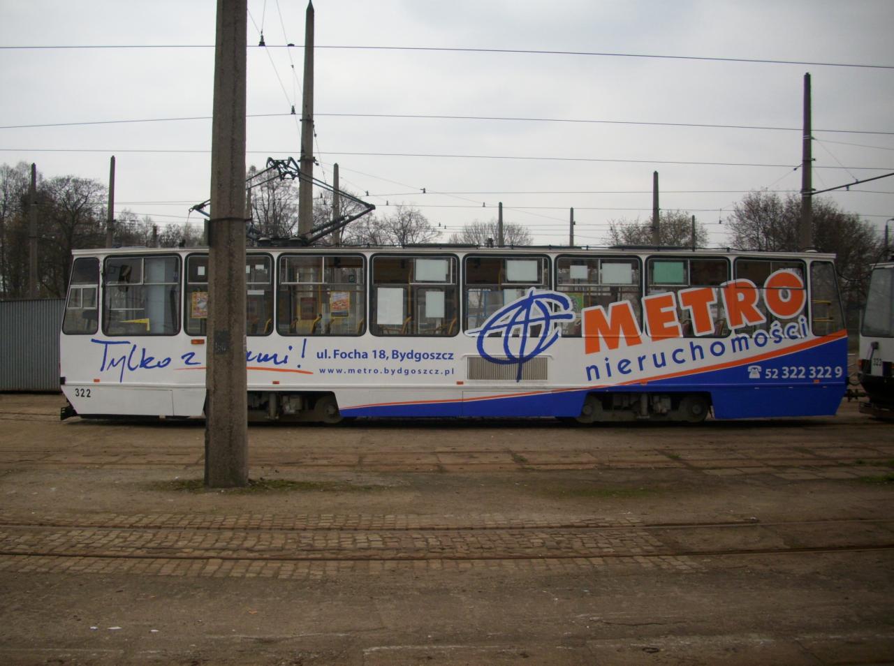 Tramwaj z reklamą firmy zajmującej się nieruchomościami, zaparkowany na terenie zajezdni, widoczne napisy reklamowe i adres firmy.
