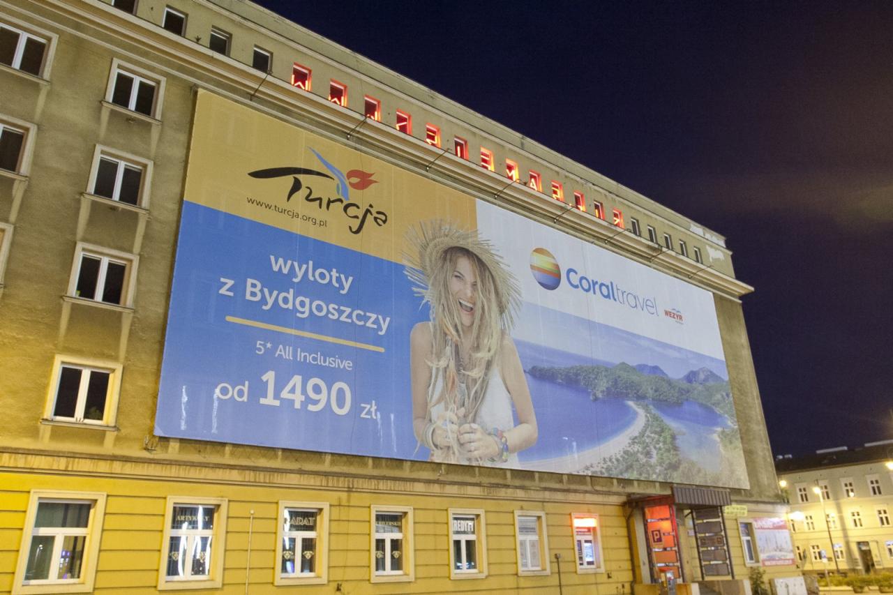 Reklama biura podróży 'Turcja' i 'Coral Travel' na budynku w nocnym oświetleniu, oferująca wycieczki z Bydgoszczy w cenie od 1490 zł.