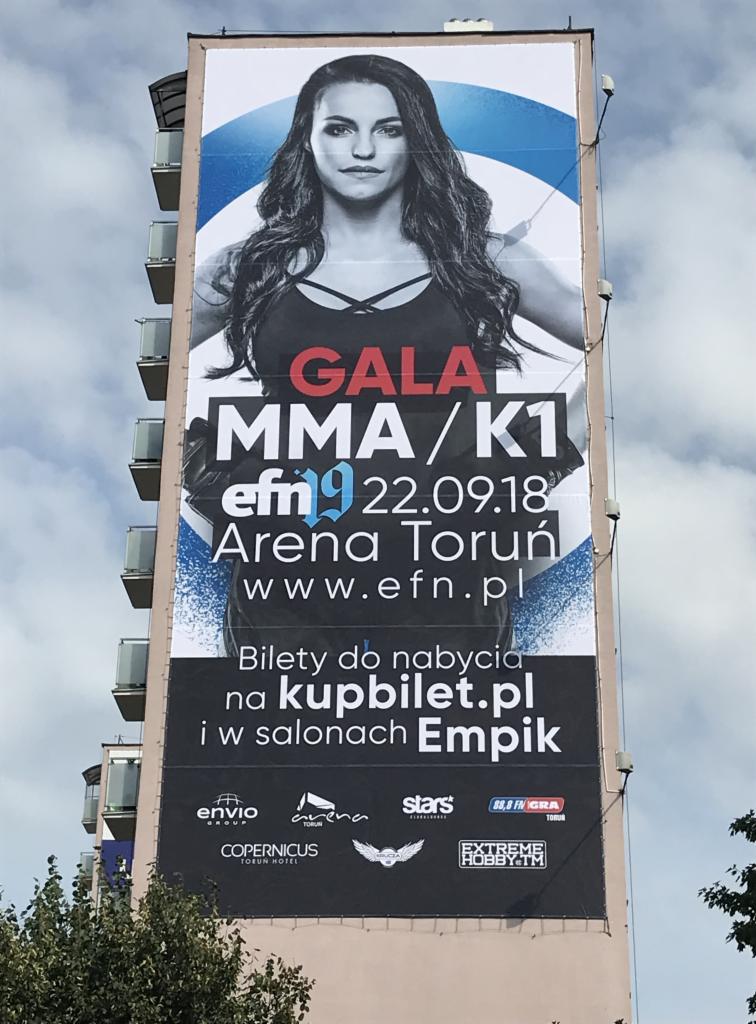 Billboard reklamujący galę MMA/K1 z wizerunkiem kobiety, umieszczony na ścianie budynku mieszkalnego. Plakat informuje o dacie i miejscu wydarzenia oraz o możliwości zakupu biletów.