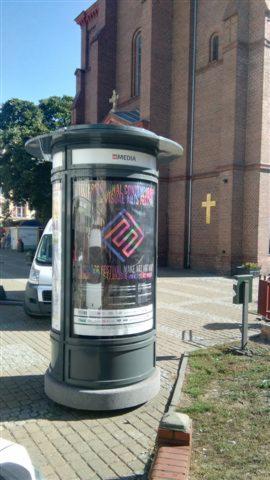 Słup ogłoszeniowy z plakatem na tle ceglanego kościoła z krzyżem, widok z poziomu chodnika.