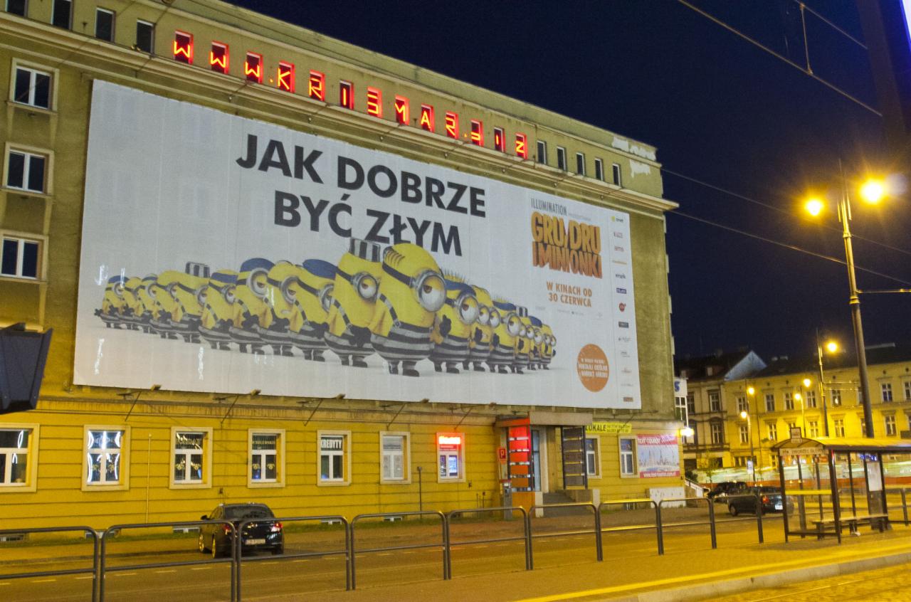 Nocny widok na budynek z dużym billboardem promującym film animowany z postaciami Minionków, oświetlony światłami ulicznymi, z widocznymi elementami miejskiej infrastruktury, takimi jak przystanek...