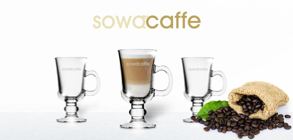 Trzy szklanki z uchwytem na nóżce, jedna wypełniona kawą latte, obok rozsypane ziarna kawy z worka jutowego, na białym tle, logo firmy u góry.