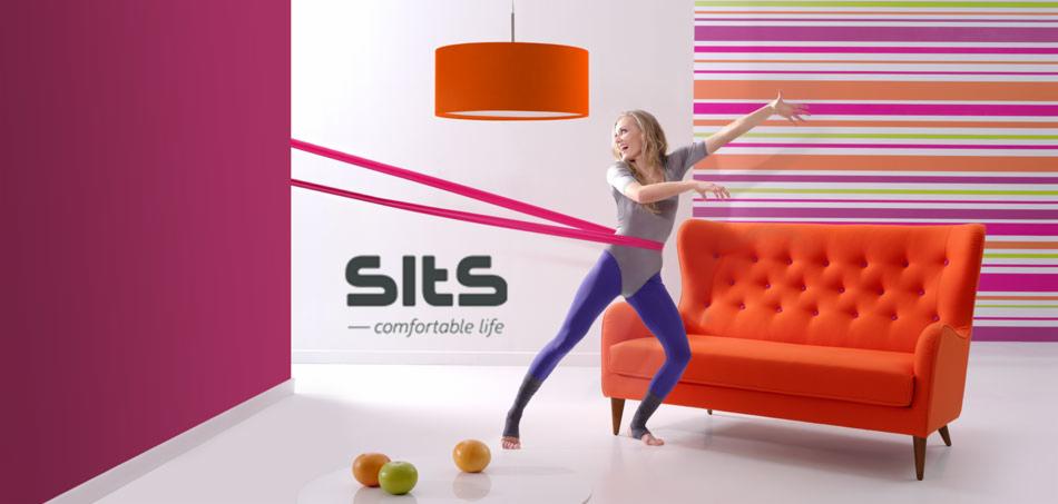 Jasne wnętrze z pomarańczową sofą, lampą i kolorową tapetą w paski. Kobieta w stroju gimnastycznym ćwiczy z różową gumą. Na podłodze leżą owoce. Widoczne logo firmy meblowej.