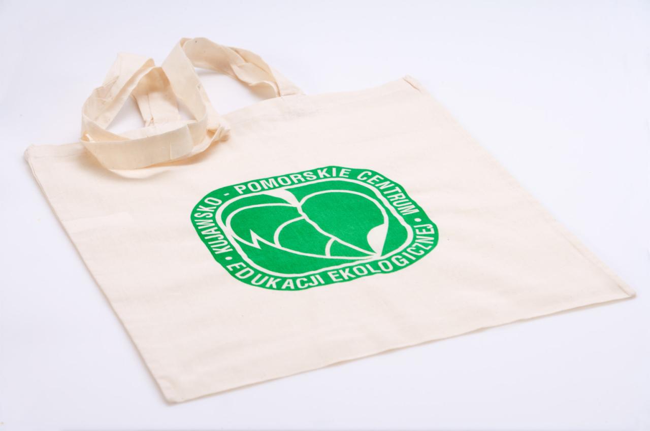 Beżowa torba bawełniana z zielonym nadrukiem logo 'Pomorskie Kujawsko Centrum Edukacji Ekologicznej' leżąca na białym tle.