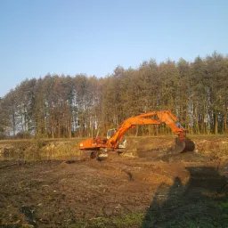 Pomarańczowa koparka w trakcie prac ziemnych na podmokłym terenie, w tle rząd drzew i błękitne niebo.