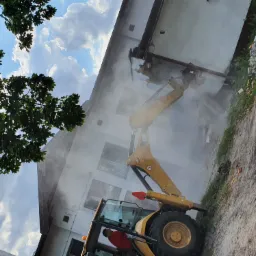 Żółta koparka CAT w trakcie kruszenia ściany budynku, unoszący się pył, widok z dołu na tle nieba z drzewami.