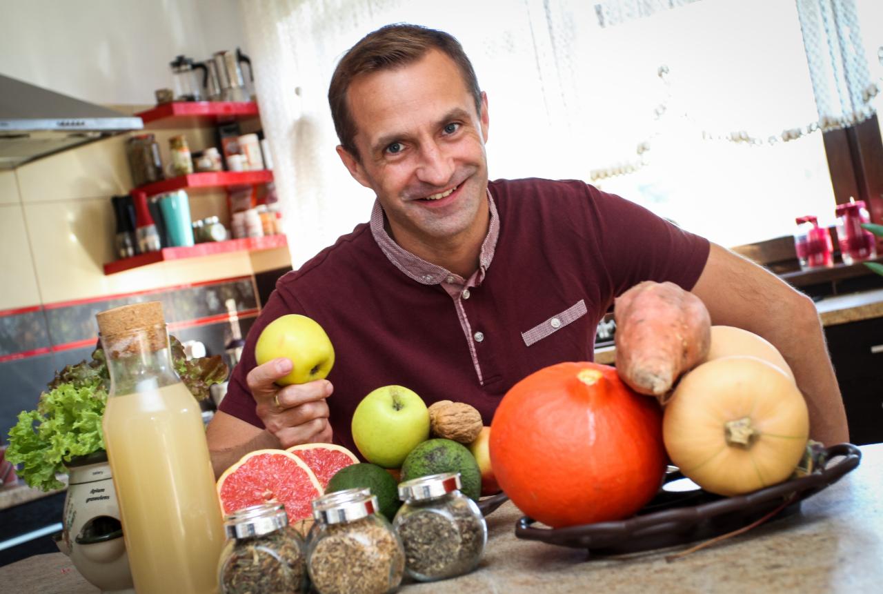 Uśmiechnięty mężczyzna w kuchni prezentuje świeże owoce i warzywa, w tym jabłka, grejpfruty, dynie i bataty, obok butelki soku i słoików z przyprawami.