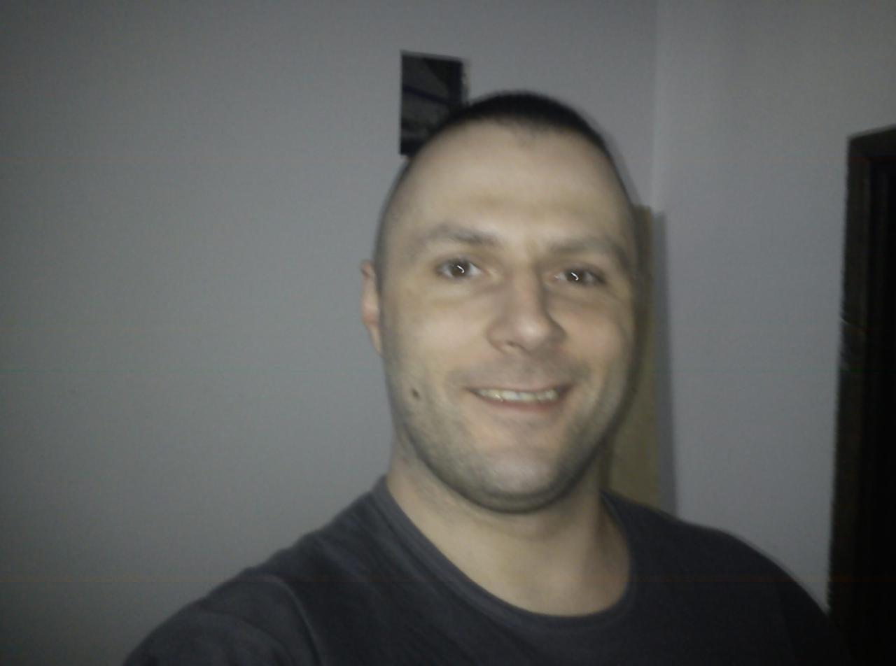 Uśmiechnięty mężczyzna z krótkimi włosami na tle ściany z otworem i fragmentem drzwi. Portret w pomieszczeniu ze słabym oświetleniem.