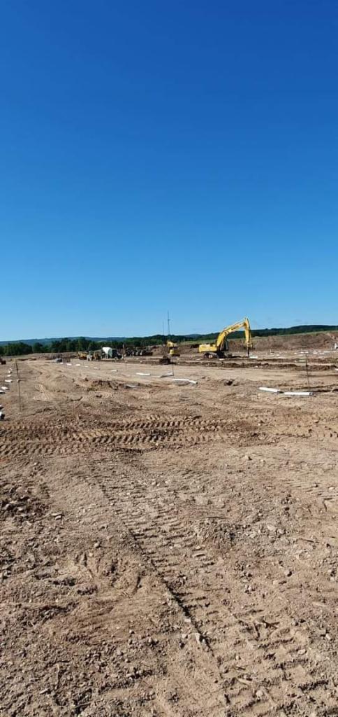 Przygotowanie terenu pod budowę z widocznymi śladami opon, żółtą koparką i innymi maszynami w oddali, błękitne niebo w tle.