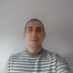 Mężczyzna w bluzie w paski na tle szarej ściany. Ujęcie od przodu, widoczna górna część ciała. Neutralny wyraz twarzy, krótko ostrzyżone włosy.