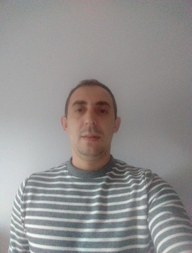 Mężczyzna w bluzie w paski na tle szarej ściany. Ujęcie od przodu, widoczna górna część ciała. Neutralny wyraz twarzy, krótko ostrzyżone włosy.