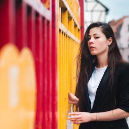 Emc² Media Piotr Skibiński - Portret kobiety w czarnej marynarce, opierającej dłoń o jaskrawo pomalowaną, metalową barierkę na tle miejskiej zabudowy.