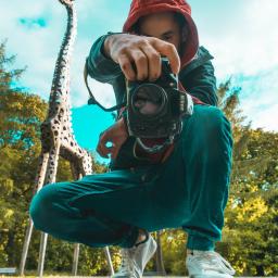 Emc² Media Piotr Skibiński - Mężczyzna w czerwonej bluzie z kapturem kuca, trzymając aparat Nikon D7100, z rzeźbą żyrafy w tle.