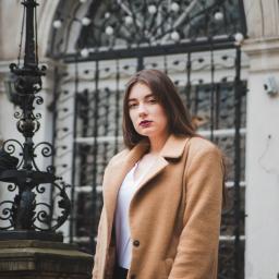 Emc² Media Piotr Skibiński - Portret kobiety w karmelowym płaszczu na tle architektonicznej bramy z kutego żelaza i kamiennych filarów.