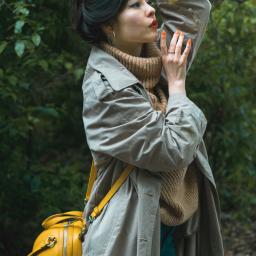 Emc² Media Piotr Skibiński - Kobieta w beżowym płaszczu i żółtej torebce na tle zieleni, dłoń z pomarańczowymi paznokciami przy głowie, usta pomalowane na czerwono.