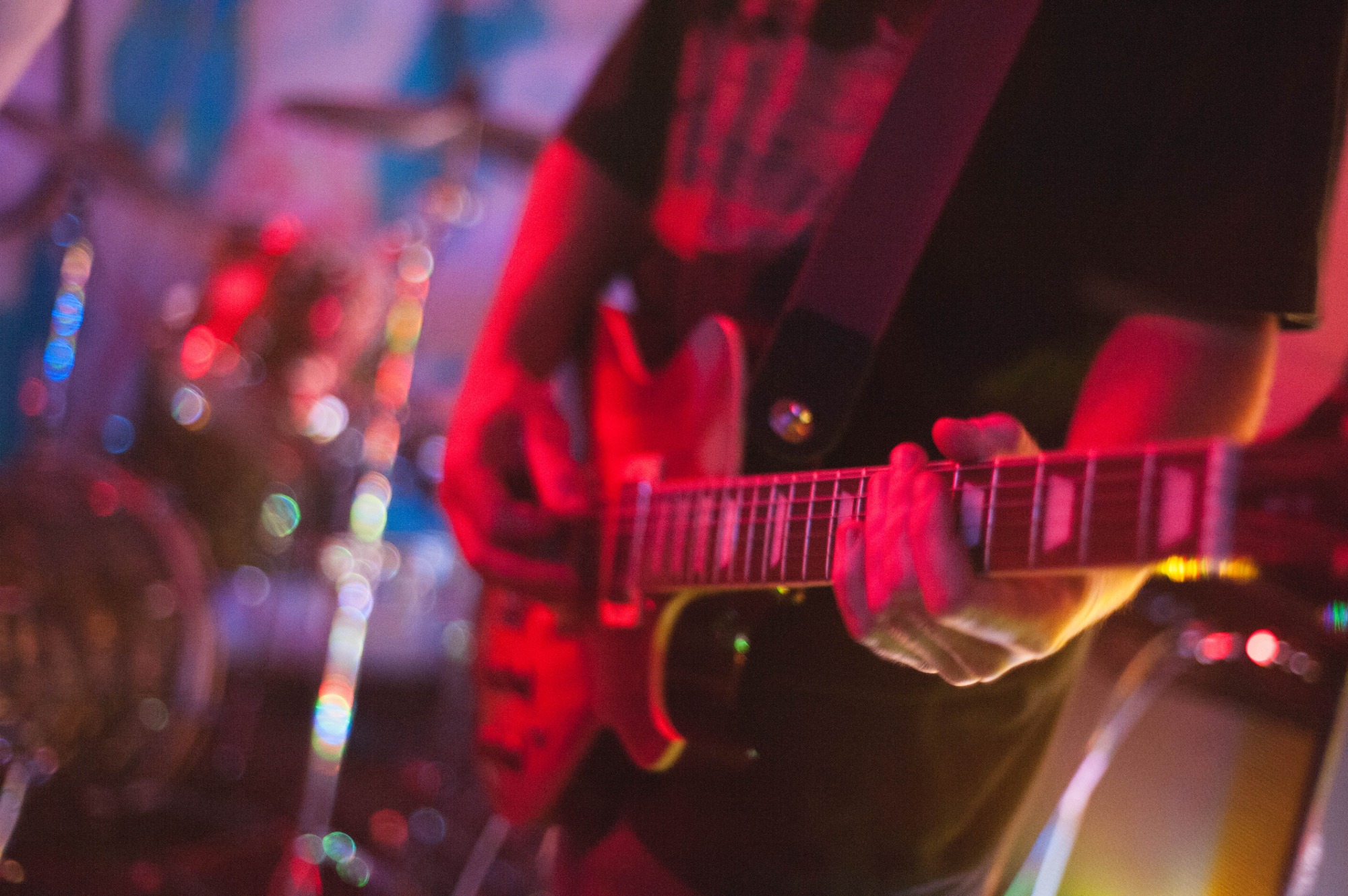 Muzyk grający na czerwonej gitarze elektrycznej na scenie, widoczne ręce na gryfie, rozmyte bębny w tle, oświetlenie sceniczne w odcieniach czerwieni i niebieskiego.