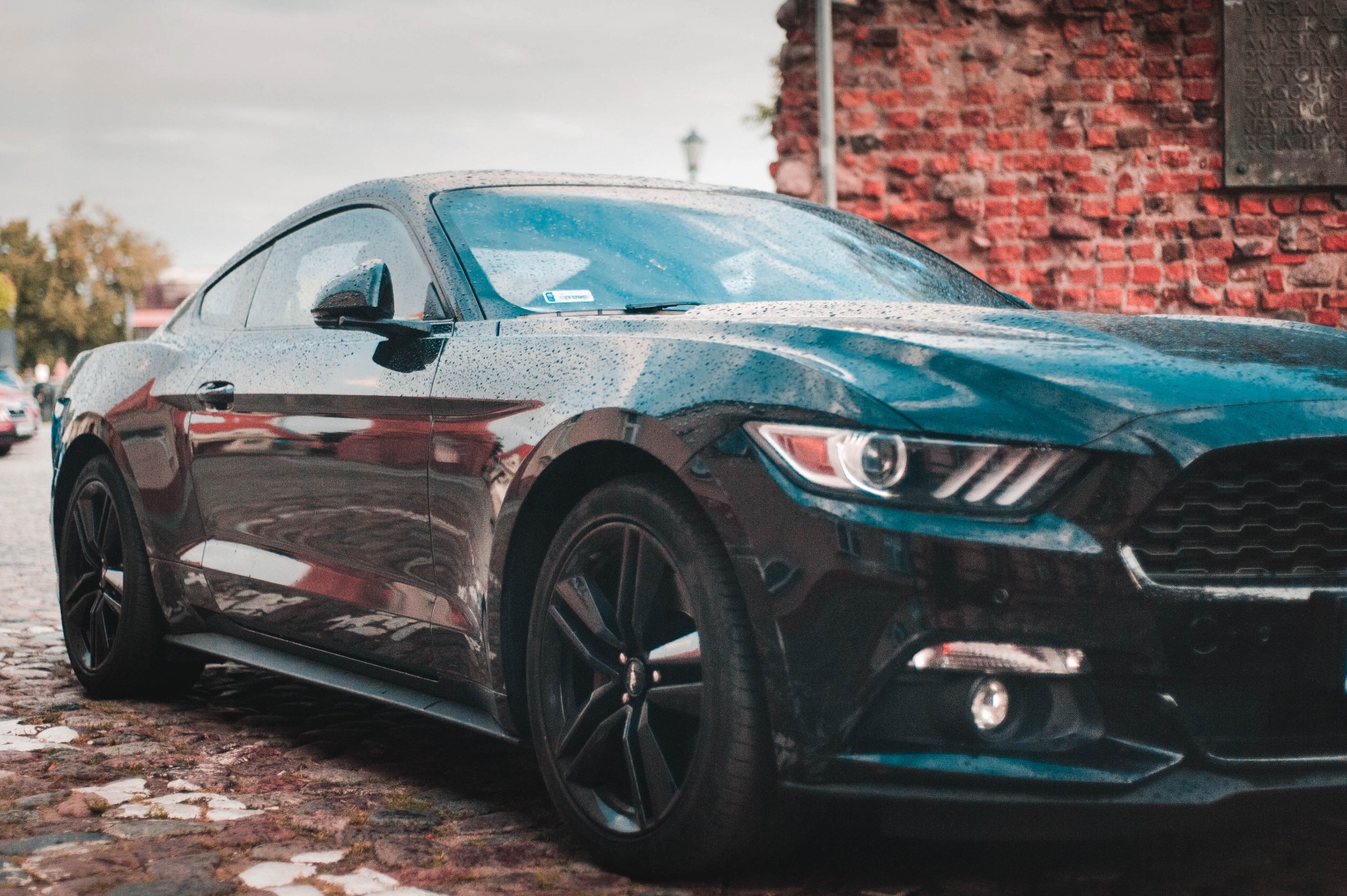 Czarny Ford Mustang, pokryty kroplami deszczu, zaparkowany na brukowanej ulicy, z ceglaną ścianą w tle.
