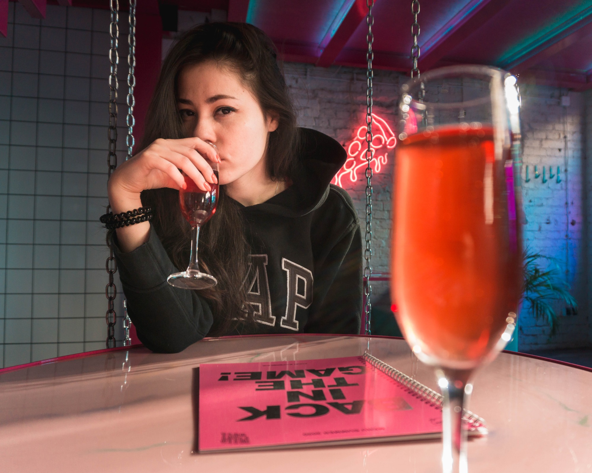 Kobieta w czarnej bluzie z kapturem pije różowy napój z kieliszka na tle neonowej pizzy i łańcuchów, z notatnikiem 'Hack the Game!' na różowym stole.