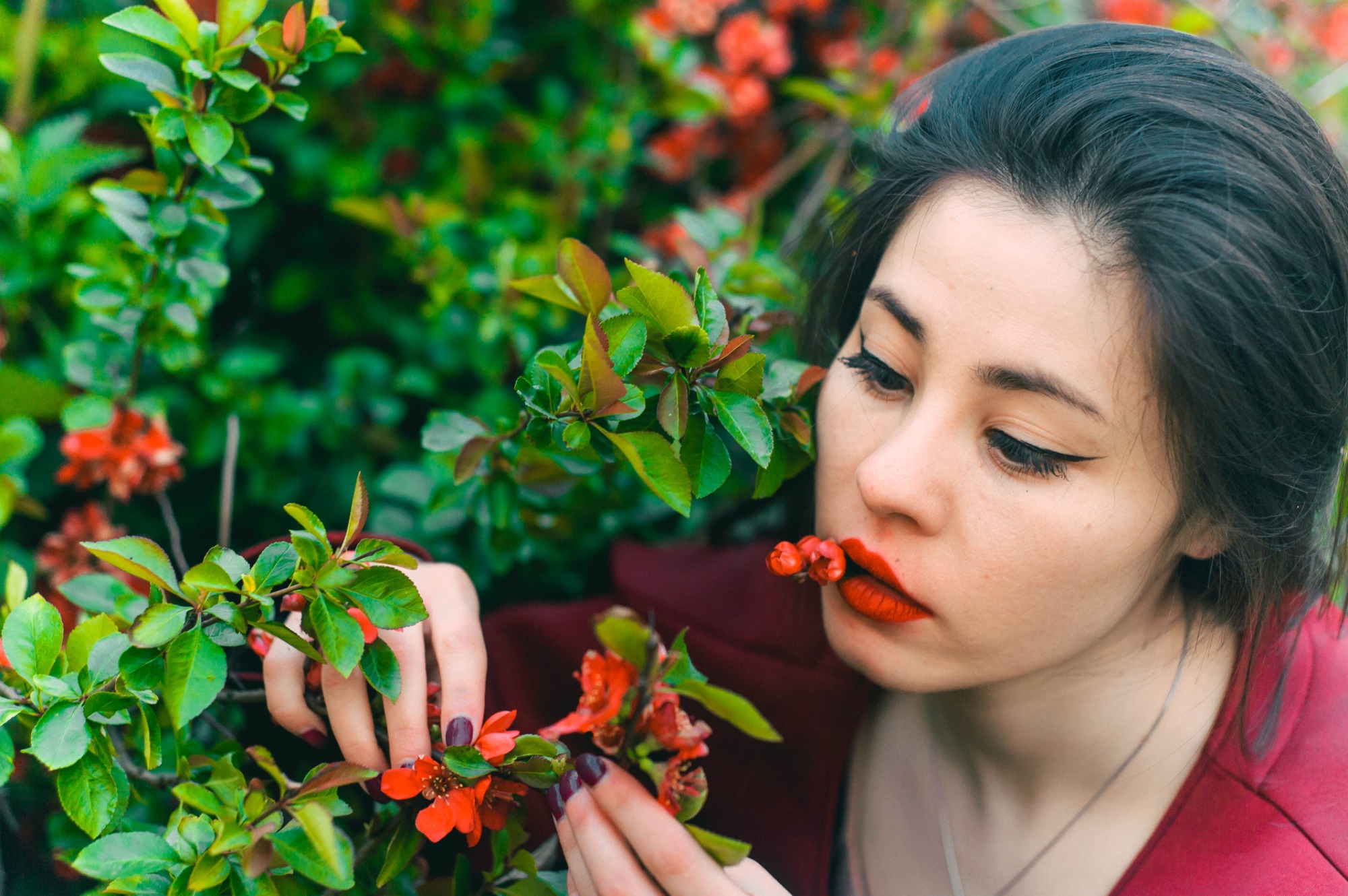 Portret kobiety z czerwonymi ustami, trzymającej kwitnącą gałązkę na tle zieleni. Uwagę przykuwa nietypowe ujęcie i intensywna kolorystyka kwiatów.