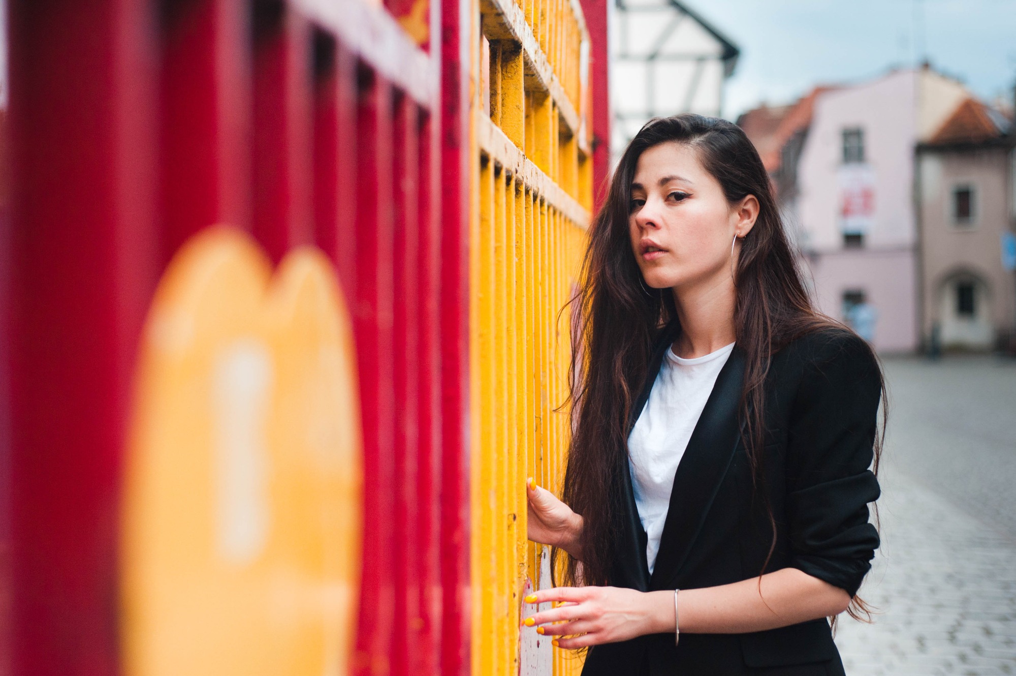 Portret kobiety w czarnej marynarce, opierającej dłoń o jaskrawo pomalowaną, metalową barierkę na tle miejskiej zabudowy.