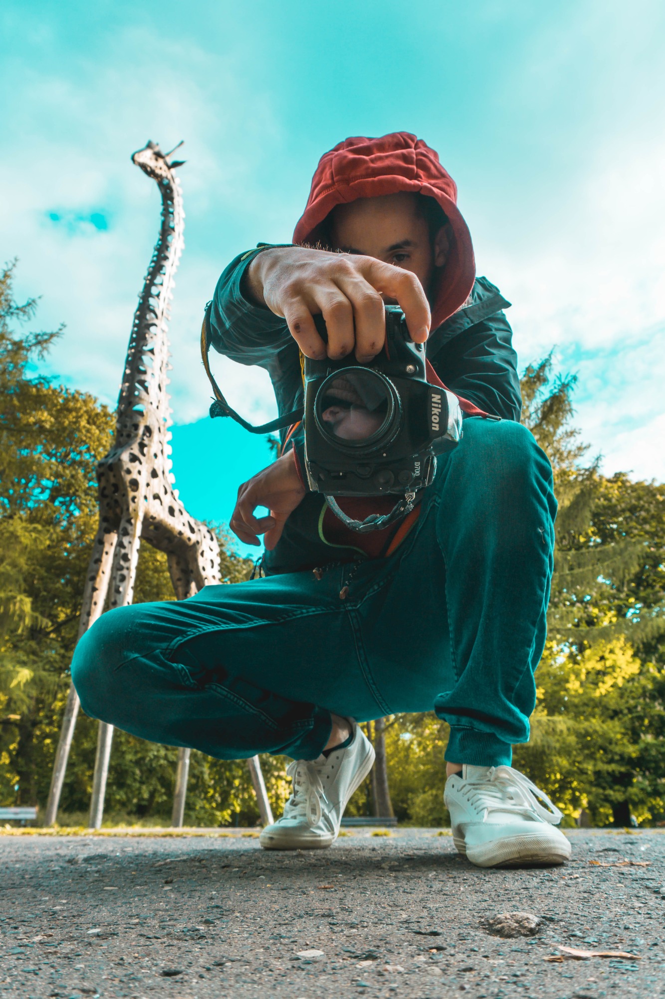 Mężczyzna w czerwonej bluzie z kapturem kuca, trzymając aparat Nikon D7100, z rzeźbą żyrafy w tle.