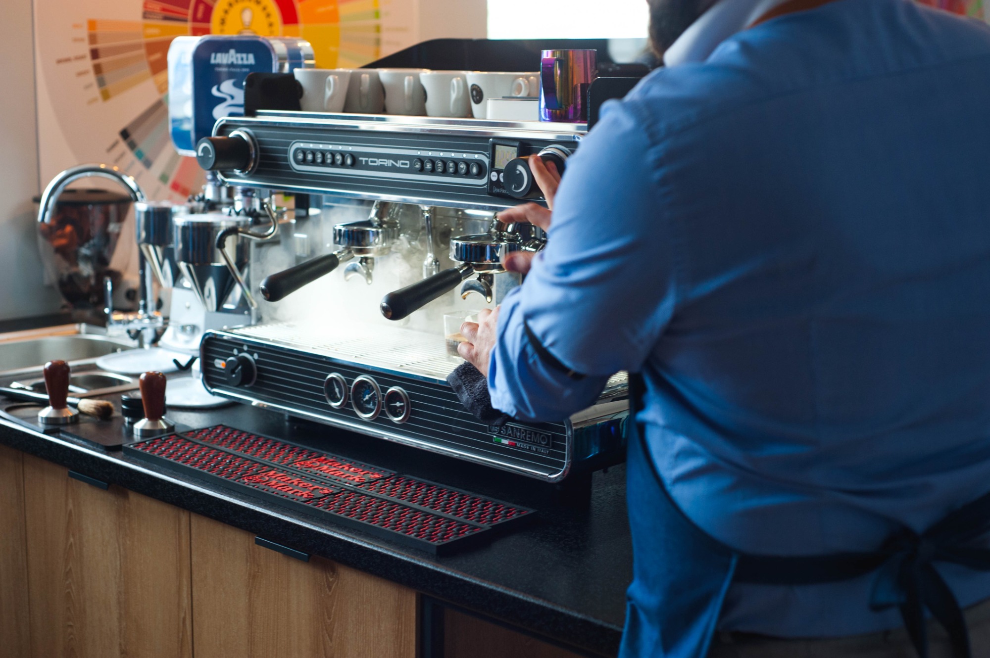 Barista w niebieskiej koszuli przygotowuje espresso na profesjonalnym ekspresie Sanremo Torino, z parą unoszącą się nad filiżanką, na tle kolorowej mapy smaków kawy Lavazza.