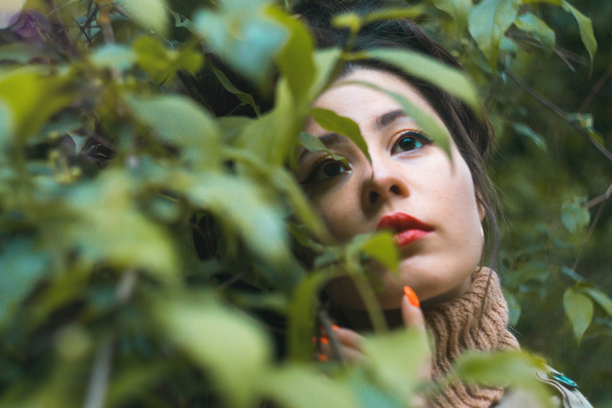 Portret kobiety z czerwonymi ustami, spoglądającej w górę, częściowo zasłoniętej zielonymi liśćmi, z pomarańczowym manicurem.