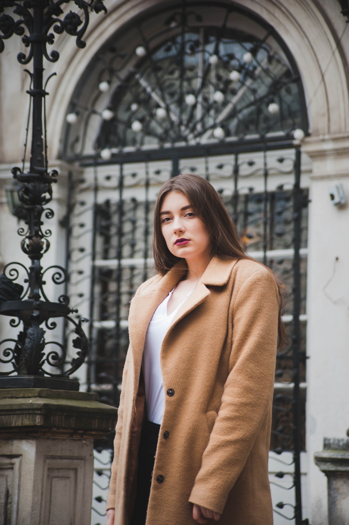Portret kobiety w karmelowym płaszczu na tle architektonicznej bramy z kutego żelaza i kamiennych filarów.