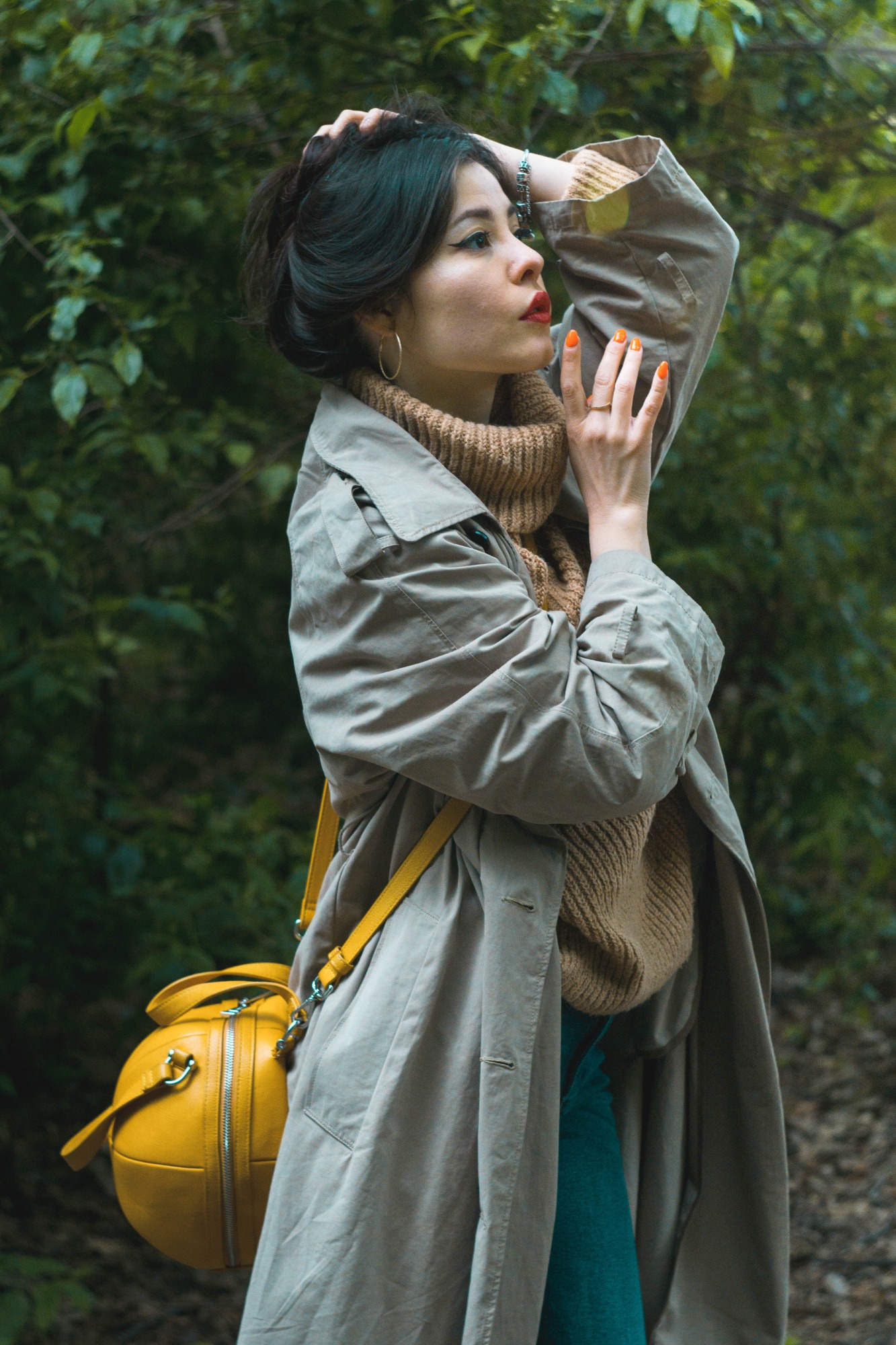 Kobieta w beżowym płaszczu i żółtej torebce na tle zieleni, dłoń z pomarańczowymi paznokciami przy głowie, usta pomalowane na czerwono.