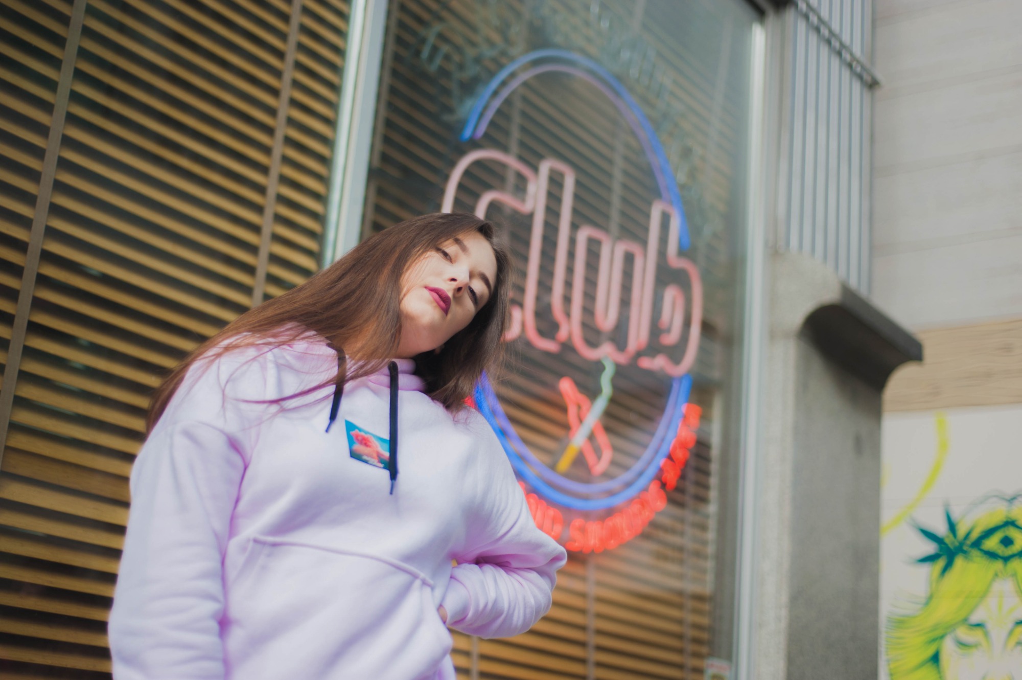 Portret młodej kobiety w jasnoróżowej bluzie z kapturem, pozującej na tle neonowego szyldu 'Club' i drewnianych żaluzji.