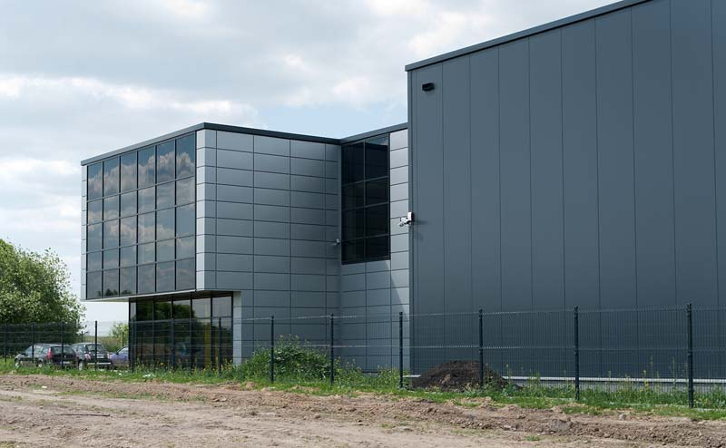 Nowoczesny budynek przemysłowy z szarych płyt warstwowych i przeszkloną częścią biurową, widoczny zza metalowego ogrodzenia, z zaparkowanymi samochodami w tle.