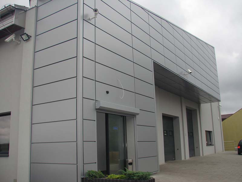 Nowoczesny budynek przemysłowy wykończony szarymi płytami elewacyjnymi, z widocznymi kamerami monitoringu i roślinami w donicach u podstawy, sfotografowany w pochmurny dzień.