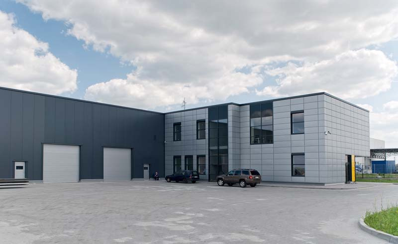 Nowoczesny budynek przemysłowy z szarymi płytami warstwowymi na elewacji i bramami rolowanymi, dwa samochody osobowe zaparkowane przed budynkiem, błękitne niebo z obłokami w tle.