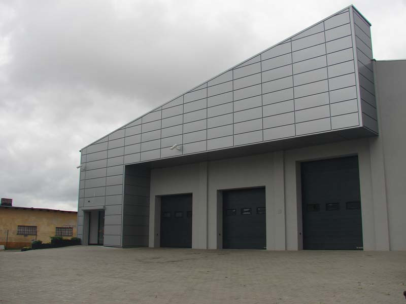 Budynek przemysłowy z elewacją z szarych płyt, trzy segmentowe bramy garażowe, widok z zewnątrz w pochmurny dzień.