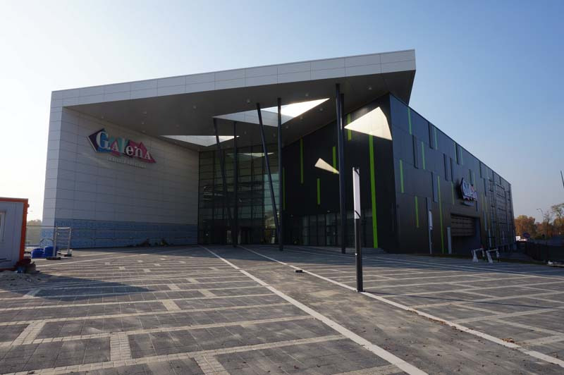 Nowoczesny budynek Galerii Handlowej Galena w Jaworznie z elewacją z płyt warstwowych w kolorach szarości i czerni, z elementami zielonymi i dużymi przeszkleniami, widziany z poziomu placu...