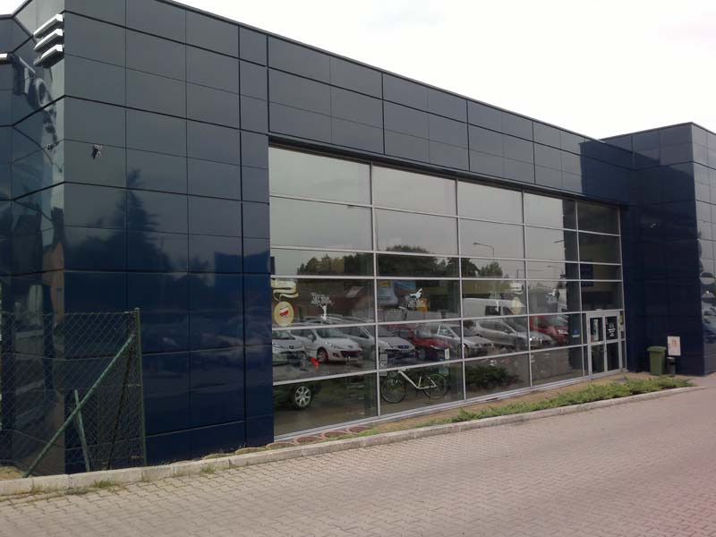 Budynek salonu samochodowego Peugeot Rybnik z elewacją z ciemnych płyt, duże okna wystawowe odbijające otoczenie, rower oparty o szybę.